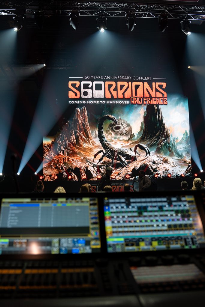 Ein Mischpult der Marke grandMA2 light ist unscharf im Vordergund, während im Hintergrund eine Bühne mit LED-Wand in der Swiss-Life-Hall in Hannover im Fokus ist. Auf der LED-Wand im Hochformat, ist das Logo der Scorpions mit den Slogan 'Coming home to Hannover', vor dem Leitmotiv für das 60-Jahre-Jubiläumskonzert der Scorpions in Hannover 2025, erkennbar. Die LED-Wand steht auf einer Bühne, mit einem langen Tisch und 12 MAC One Beam-Washlights, sowie 2 Kling und Freitag-Lautsprechern. Über der Bühne hängen Movinglights an Traversen, vor der Bühne sitzen Pressevertreter.