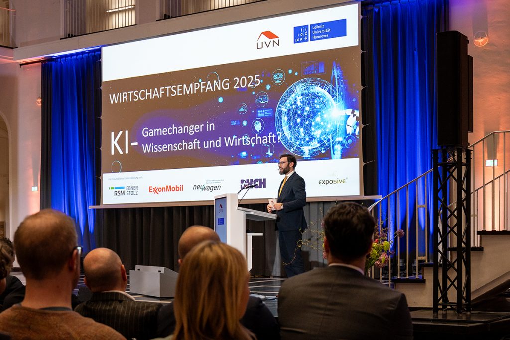 Nahaufnahme im Lichthof der Leibniz Universität Hannover: Bühne mit Leinwand „Wirtschaftsempfang 2025: KI – Gamechanger in Wissenschaft und Wirtschaft?“ und Benedikt Hüppe Hauptgeschäftsführer der UVN) am Pult, vor voll besetztem Saal.