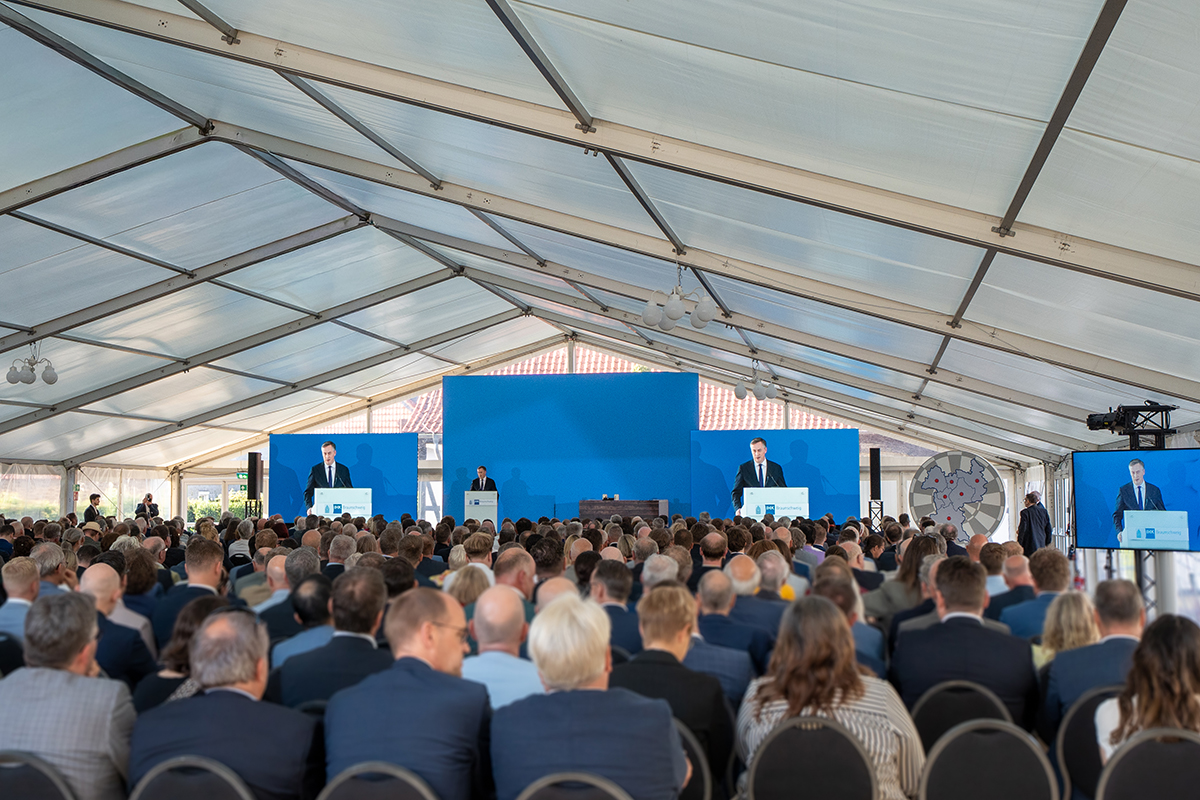 Vollbesetztes Festzelt beim IHK Sommerempfang 2025: Keynote-Speaker David McAllisteram Pult vor großem blauem Bühnen-Backdrop, links und rechts zeigen LED-Displays das Livebild; im Vordergrund dichtes Publikum, unter dem Zeltdach kugelförmige Leuchten und seitliche Kamera-/PA-Positionen.