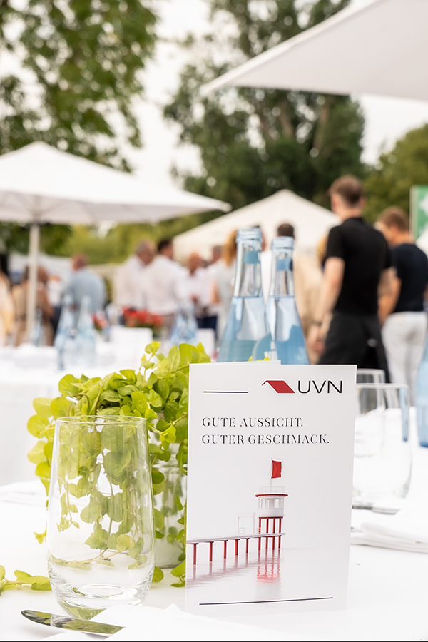 Weiß eingedeckter Tisch beim INW Sommerfest 2025 im Freien: im Vordergrund Menükarte und Branding-Karte mit UVN-Logo und dem Text „GUTE AUSSICHT. GUTER GESCHMACK.“, daneben Wassergläser, Besteck, blaue Flaschen und grüne Tischdeko; im Hintergrund Gäste unter weißen Sonnenschirmen.